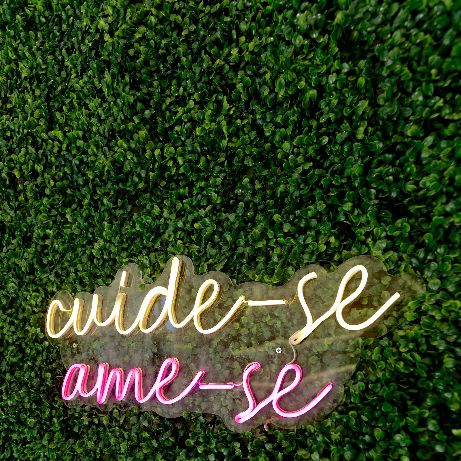 Parede de folhagem verde do Centro Estético com letreiro de neon cuide-se em luz quente e ame-se em rosa