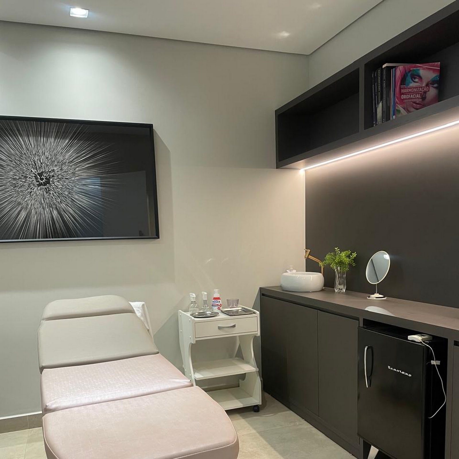 Sala de atendimento do Centro Estético com maca rosê, carrinho de produtos, pia e espelho de mão
